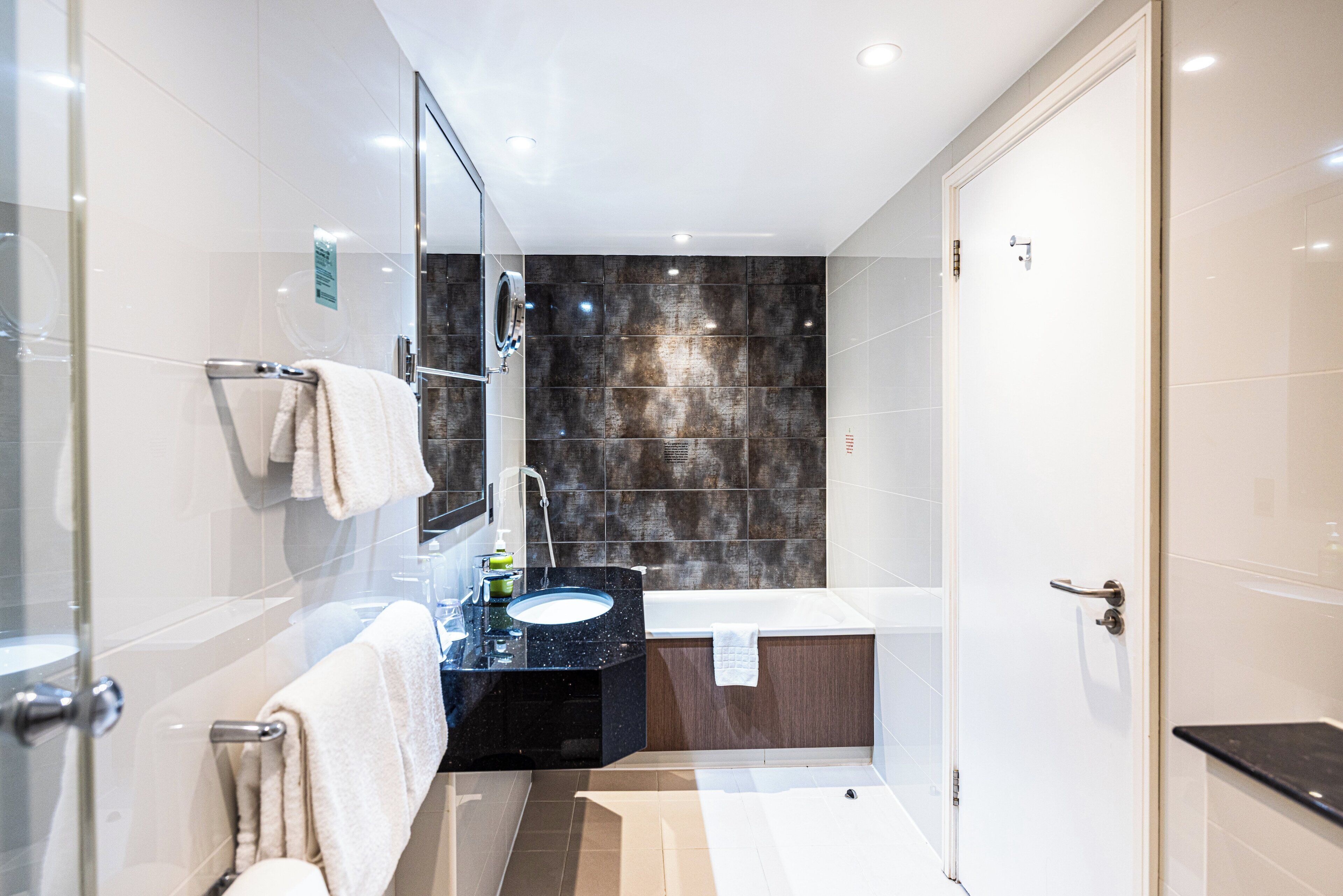 Un baño moderno con cabina de ducha de vidrio, un lavamanos negro, una bañera blanca y una pared revestida con baldosas oscuras.