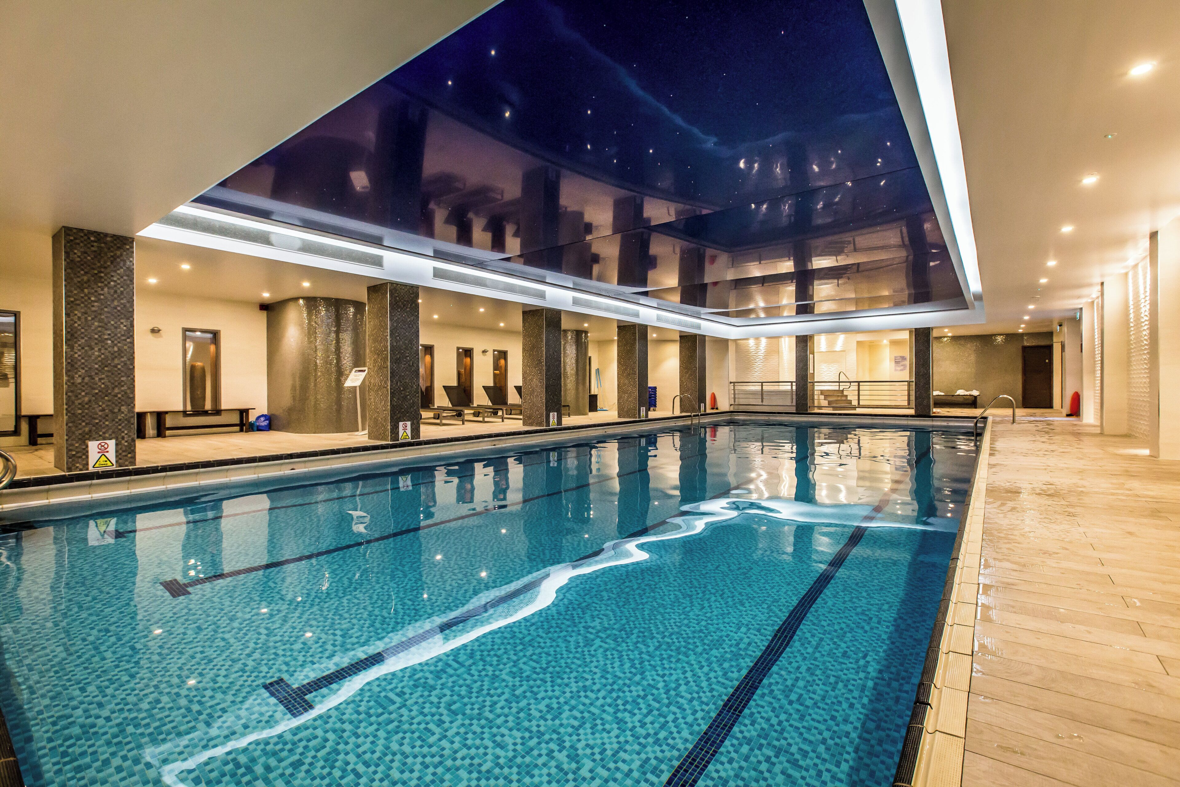 An indoor swimming pool with clear water, tiled floor, and a view of the night sky through a large window.
