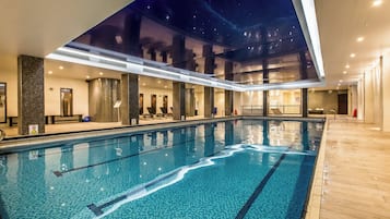 An indoor swimming pool with clear water, tiled floor, and a view of the night sky through a large window.