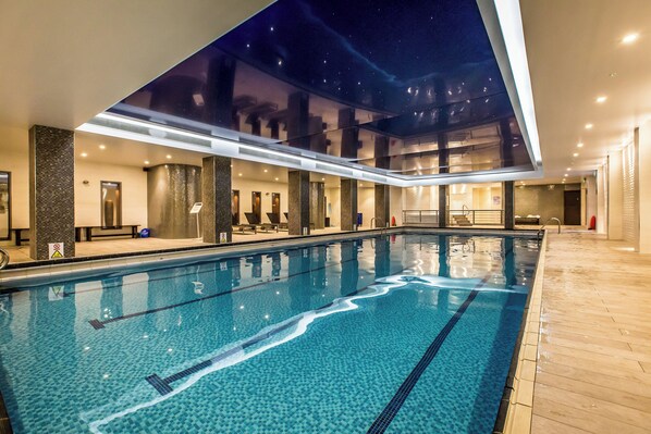 An indoor swimming pool with clear water, tiled floor, and a view of the night sky through a large window.