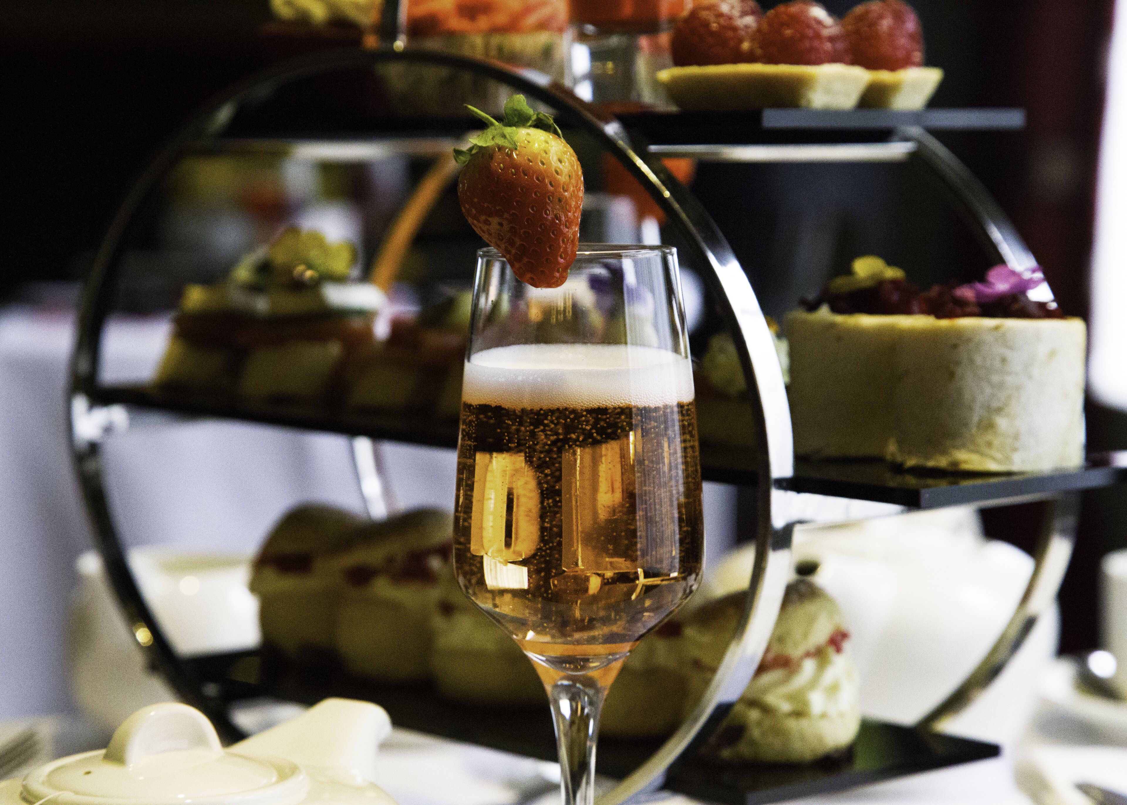 A glass of beer with a strawberry garnish, a dessert trolley with various sweets, and a cake in the background.