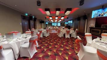 A banquet hall with round tables, white chairs with orange sashes, and a red carpeted floor.