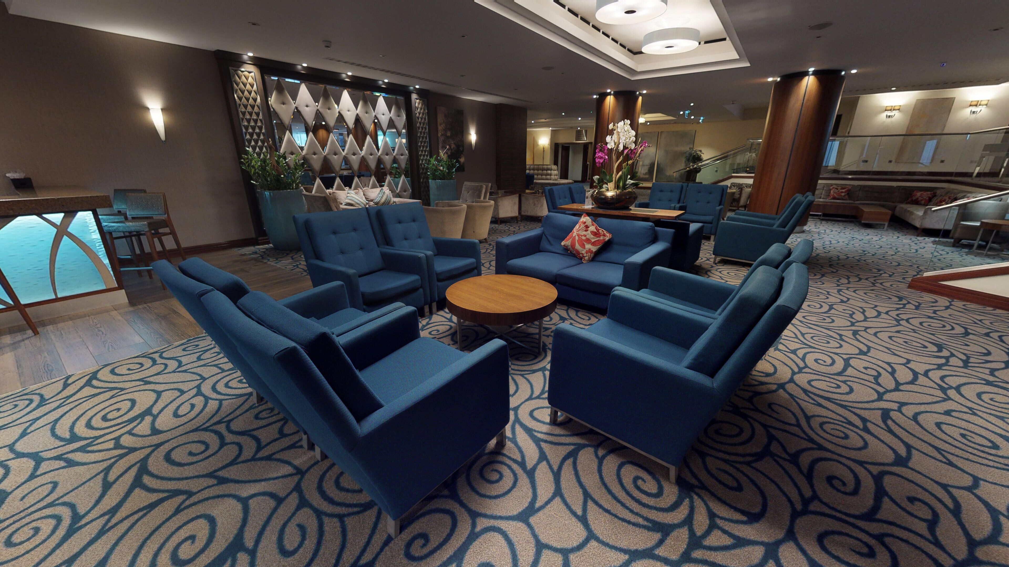 Eine Lobby mit blauen Sesseln, die um einen zentralen Couchtisch angeordnet sind, einem gemusterten Teppich und einer Wand mit geometrischen Mustern.
