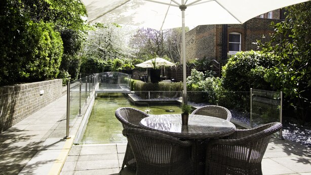A patio with wicker furniture and a large umbrella, surrounded by greenery and a brick building.