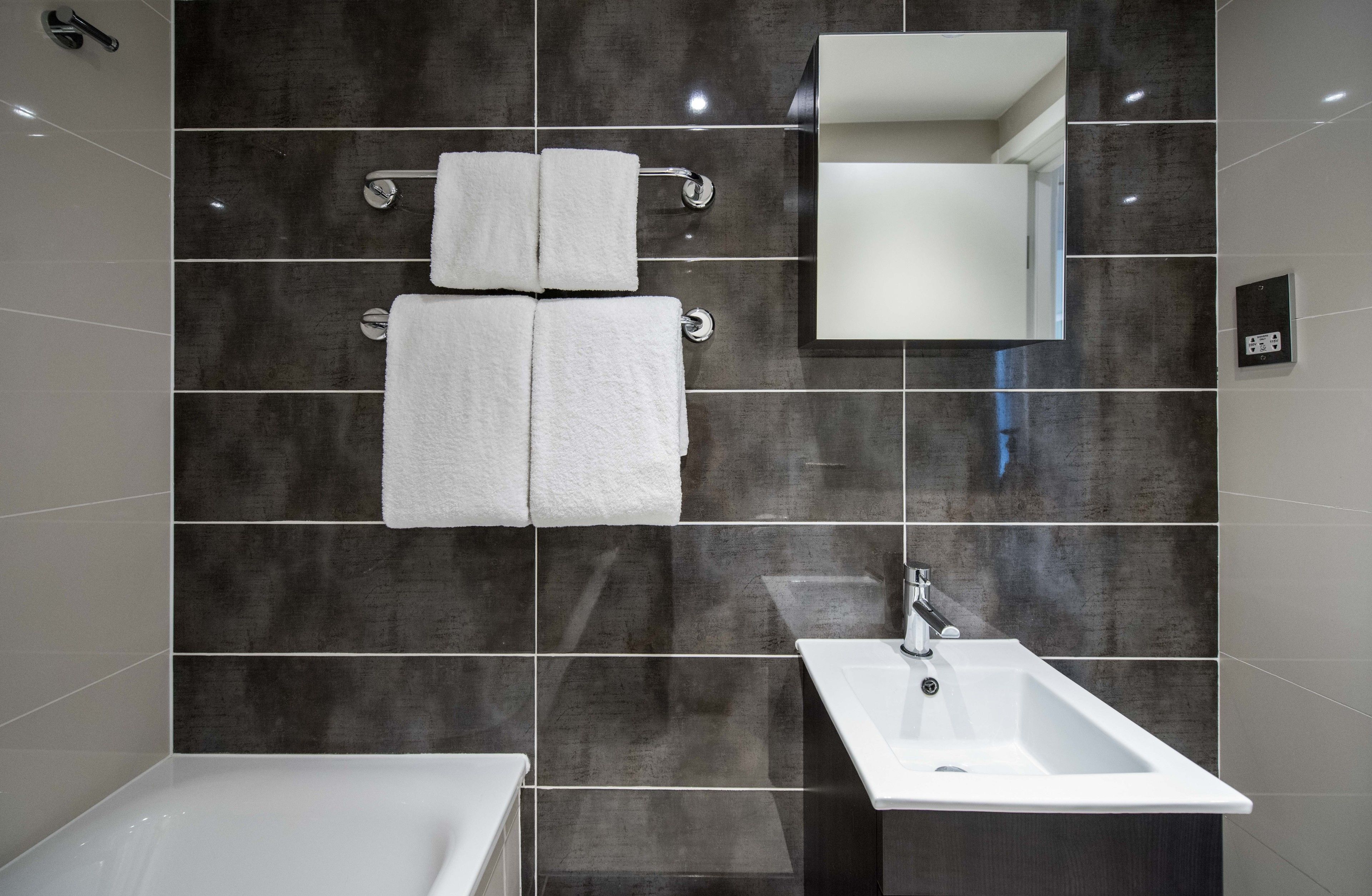 Ein modernes Badezimmer mit dunkel gefliesten Wänden, einem weißen Waschbecken und zwei weißen Handtüchern an einem Handtuchhalter.