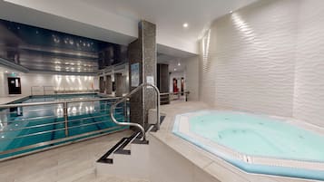 An indoor swimming pool with a separate hot tub area, a staircase leading to a higher level, and a wall with a textured pattern.
