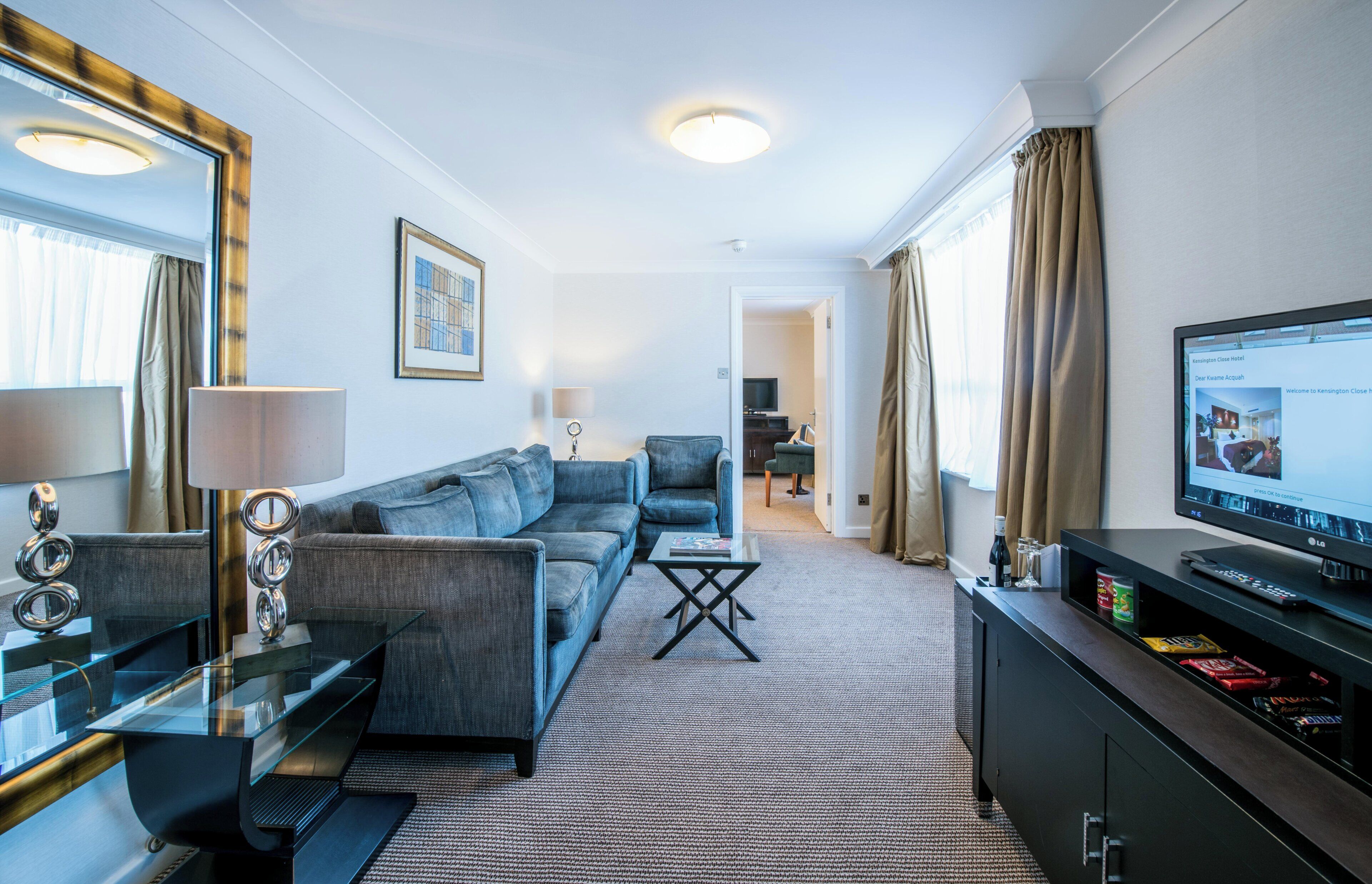 A modern hotel room with a sofa, a coffee table, a TV, and a minibar.