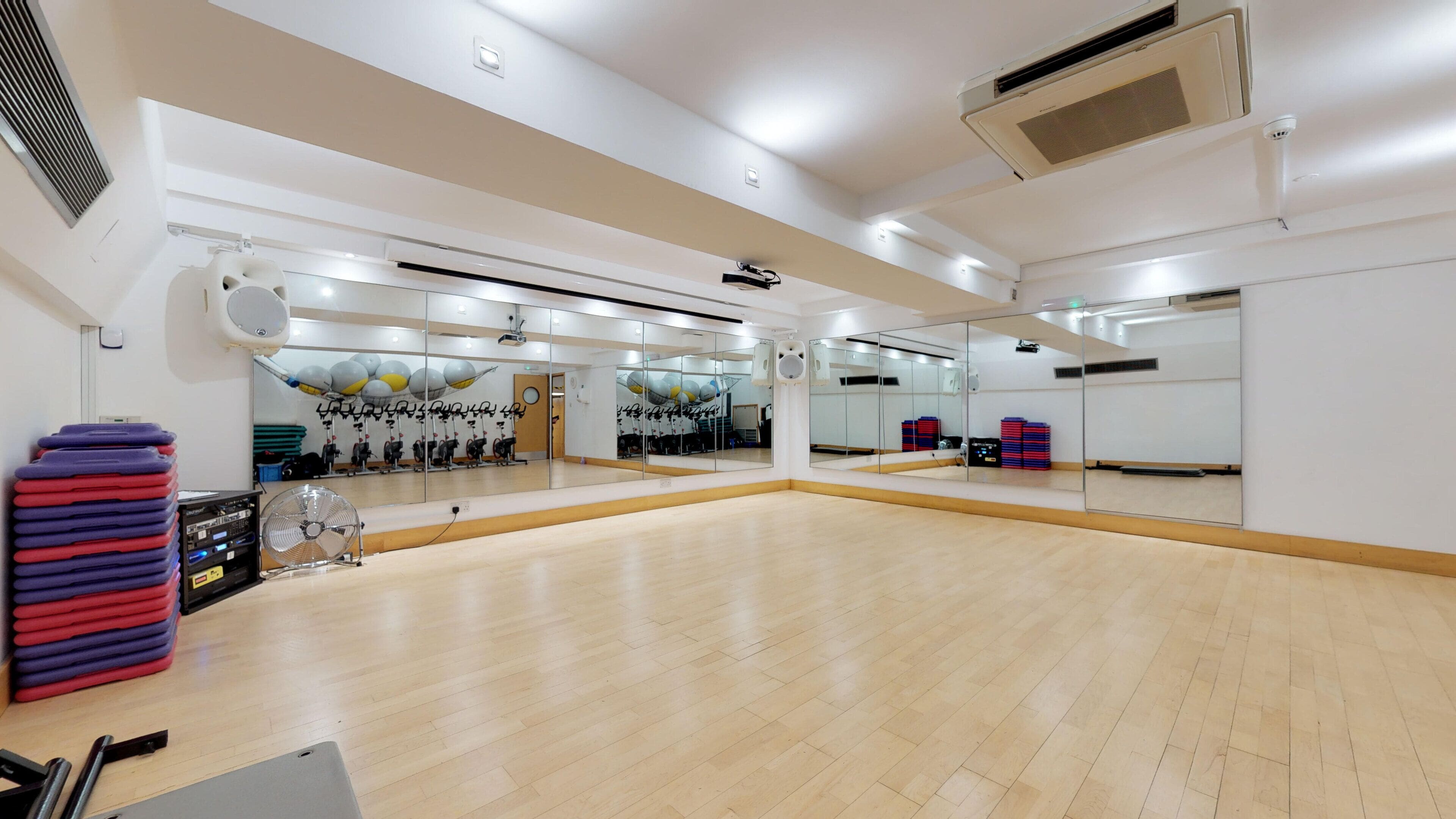 A spacious dance studio with mirrored walls, a wooden floor, and a ceiling-mounted projector.