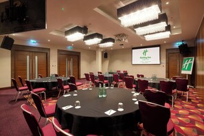 Una sala conferenze con tavoli rotondi, sedie rosse e un grande schermo con il logo Holiday Inn.