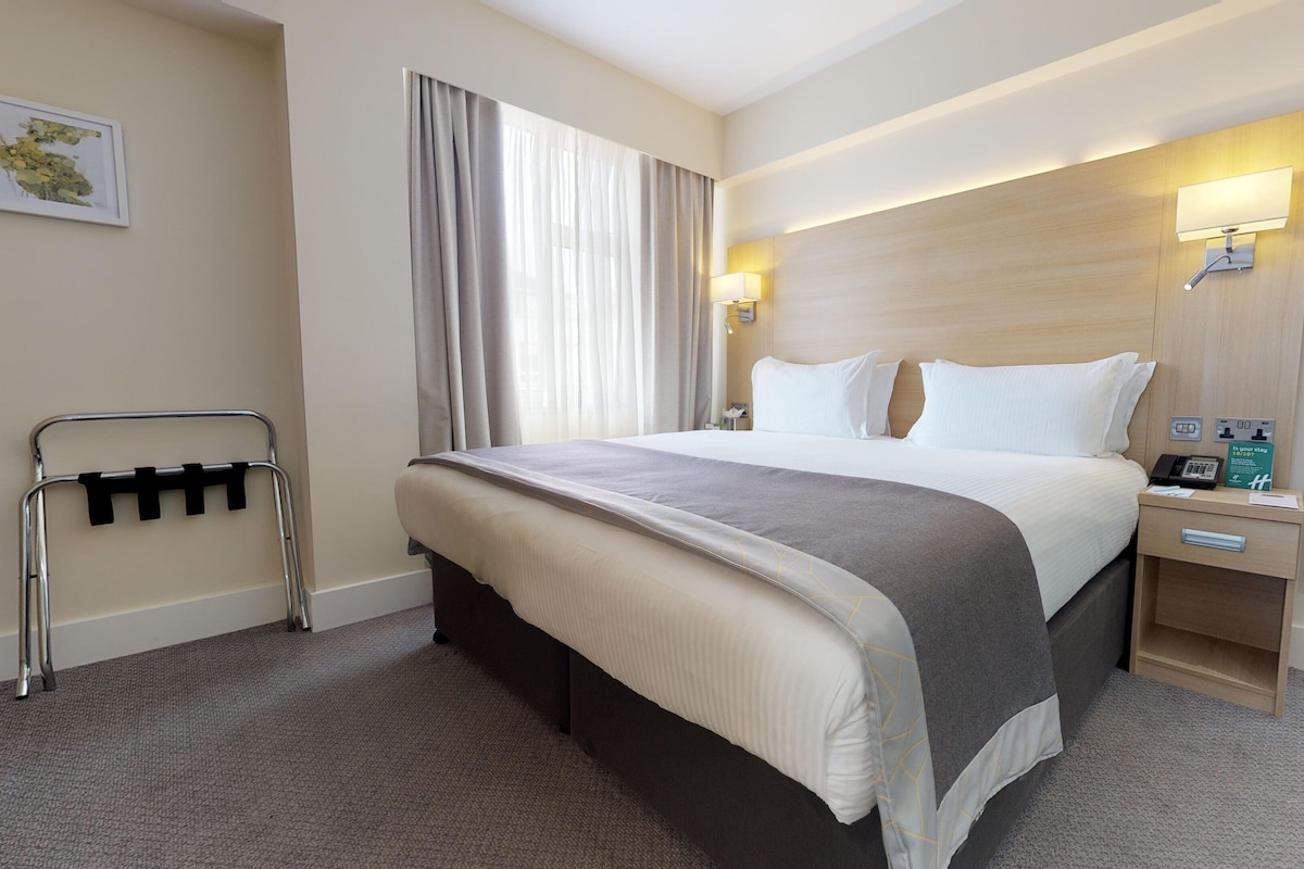 Quarto de hotel com uma cama grande, uma mesinha de cabeceira com um telefone e um aviso, uma TV em uma prateleira de madeira e uma janela com cortinas.