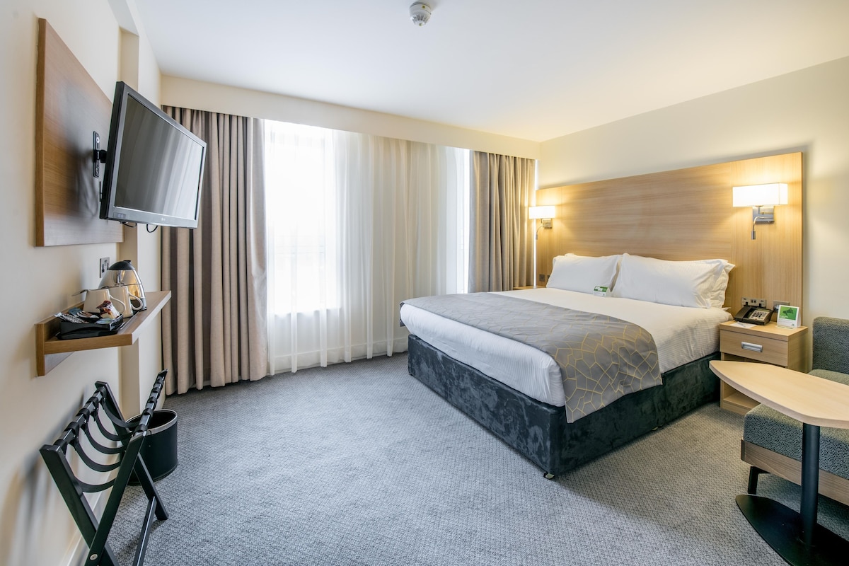 Quarto de hotel com cama, mesa de cabeceira, abajur, TV fixada na parede e uma cadeira.