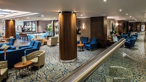 A spacious lobby with a central column, blue and beige seating, a bar area, and a patterned carpet. - Holiday Inn London - Kensington High St. by IHG (London)