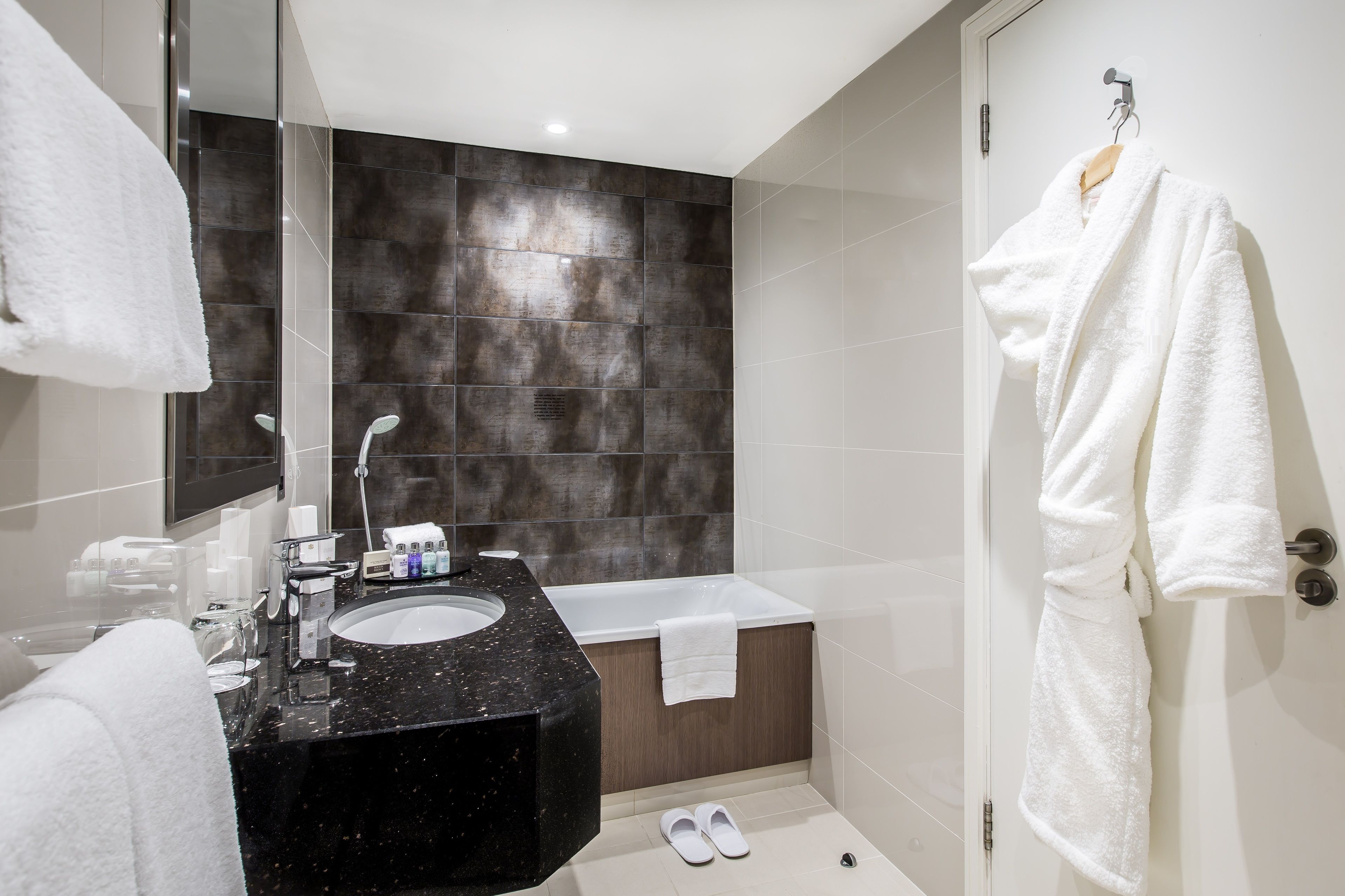 A modern bathroom with a black marble sink, a white bathtub, and a white robe hanging on the door.