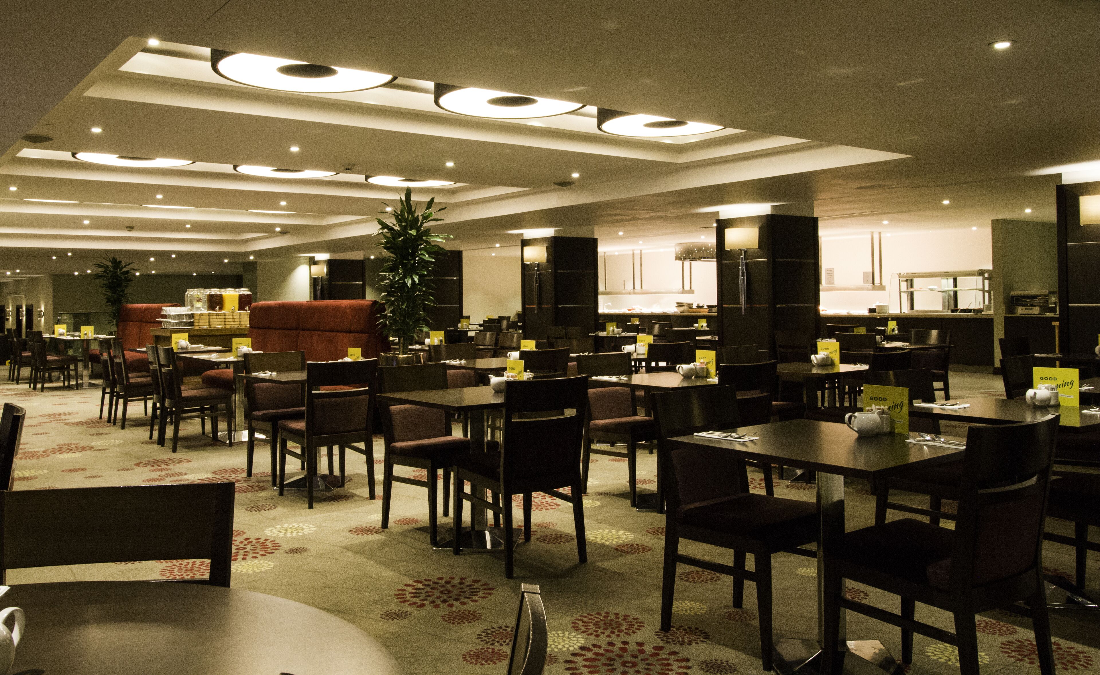 A modern dining area with multiple tables and chairs, overhead lighting, and a central plant.