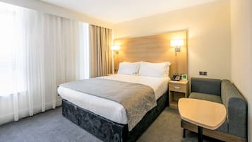 Una habitación de hotel con cama, mesita de noche, lámpara y un pequeño rincón de descanso.