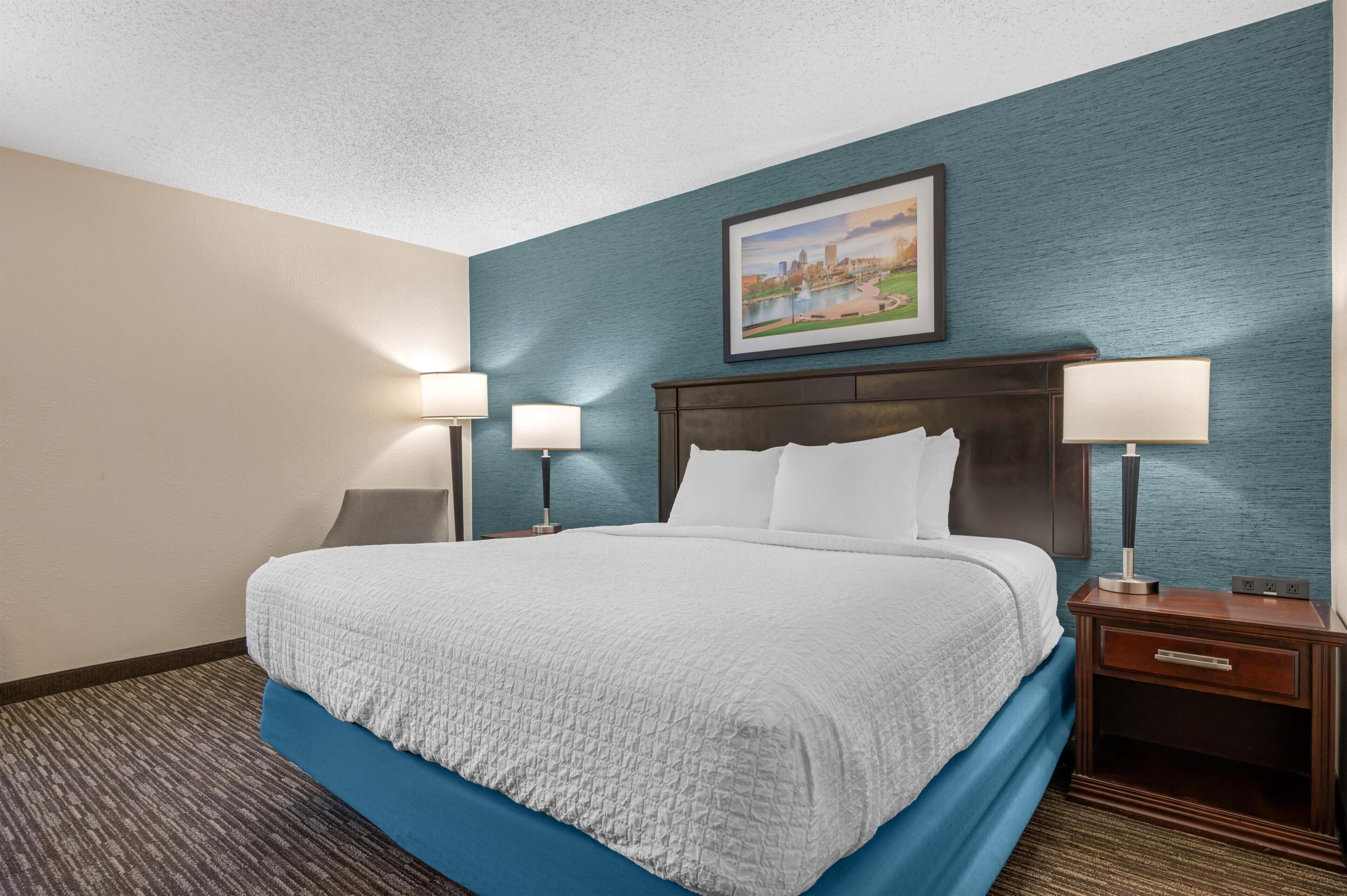 A hotel room with a bed, two bedside lamps, a nightstand, a framed picture on the wall, and a carpeted floor.