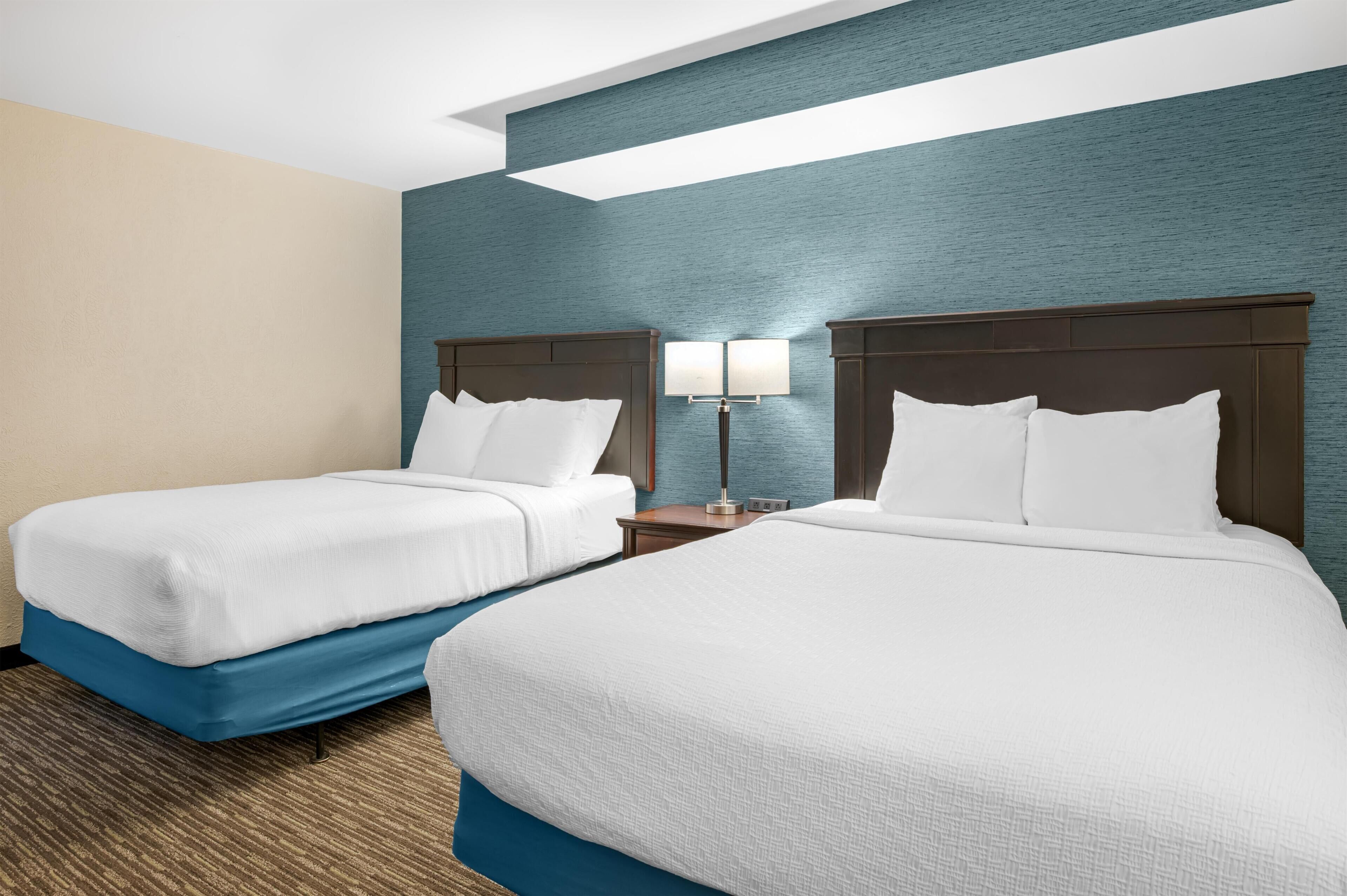 Une chambre d’hôtel avec deux lits, une table de chevet avec une lampe et un mur bleu à la texture particulière.