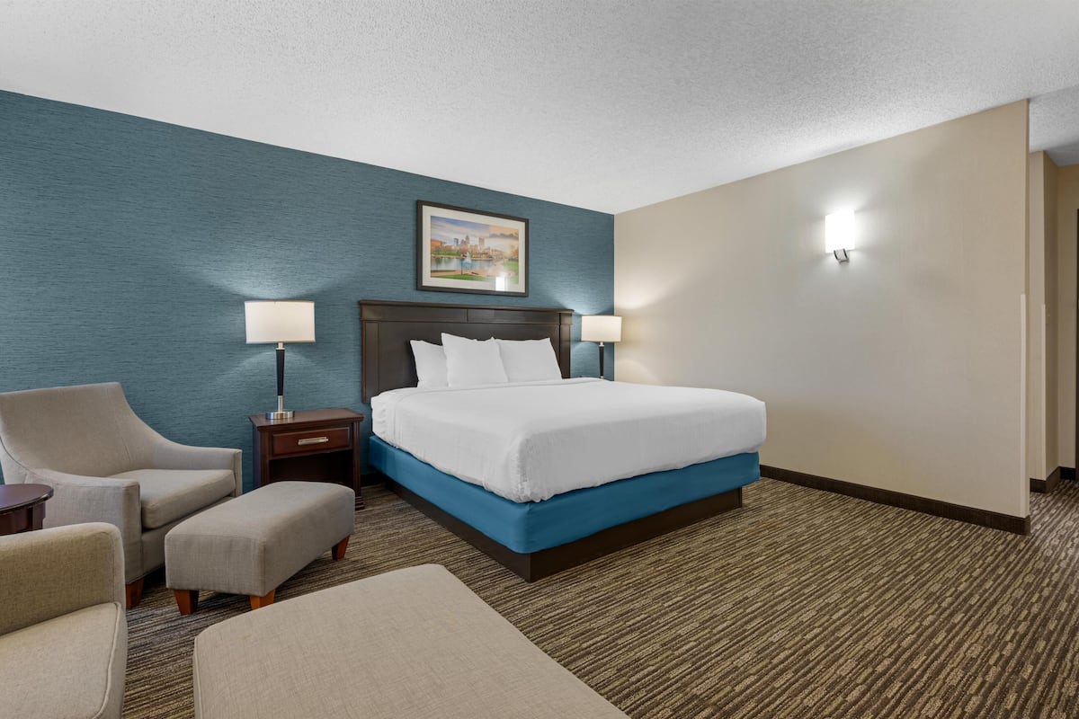 A hotel room with a bed, two bedside tables with lamps, a chair, an ottoman, and a painting on the wall.
