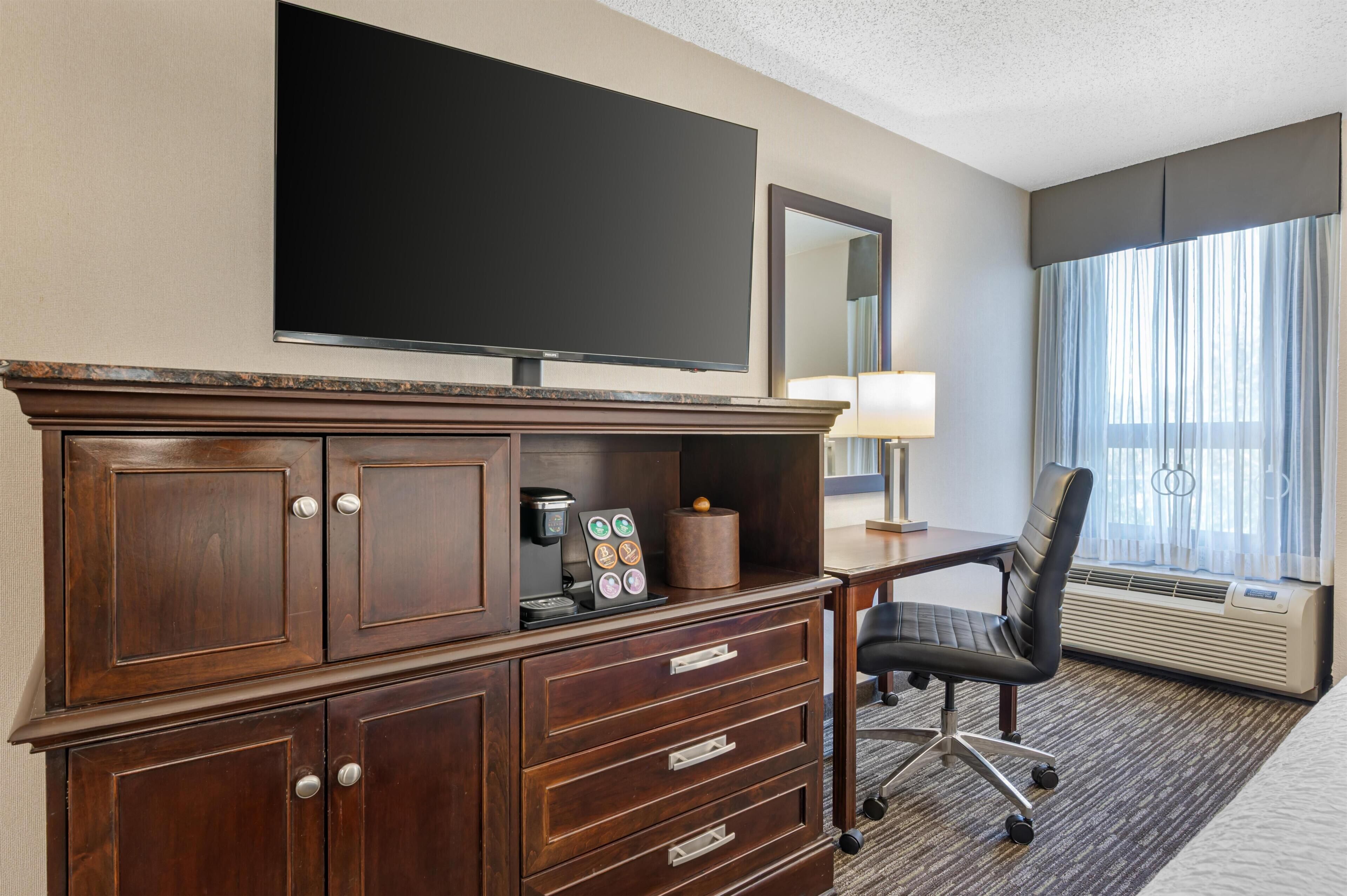 Une chambre d’hôtel équipée d’un téléviseur à écran plat, d’un bureau avec une lampe, d’une chaise, d’une machine à café et d’une grande fenêtre avec des rideaux.