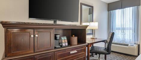 Une chambre d’hôtel équipée d’un téléviseur à écran plat, d’un bureau avec une lampe, d’une chaise, d’une machine à café et d’une grande fenêtre avec des rideaux.