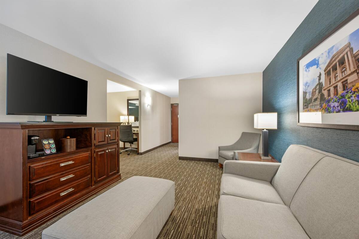 A hotel room with a flat-screen TV, a wooden cabinet, a sofa, a chair, a desk, and a framed picture on the wall.