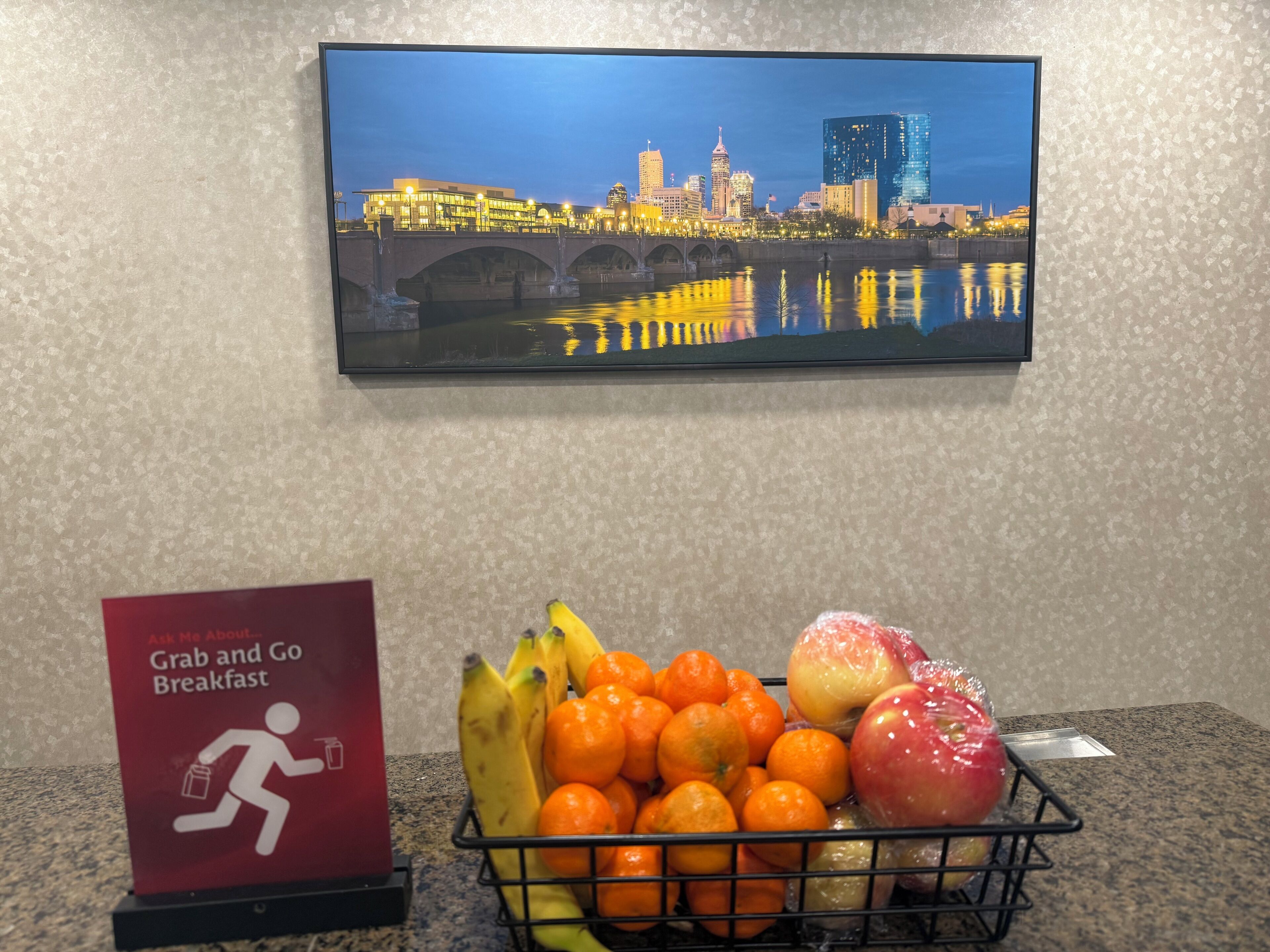A framed cityscape view, a 'Grab and Go Breakfast' sign, and a fruit basket on a countertop.