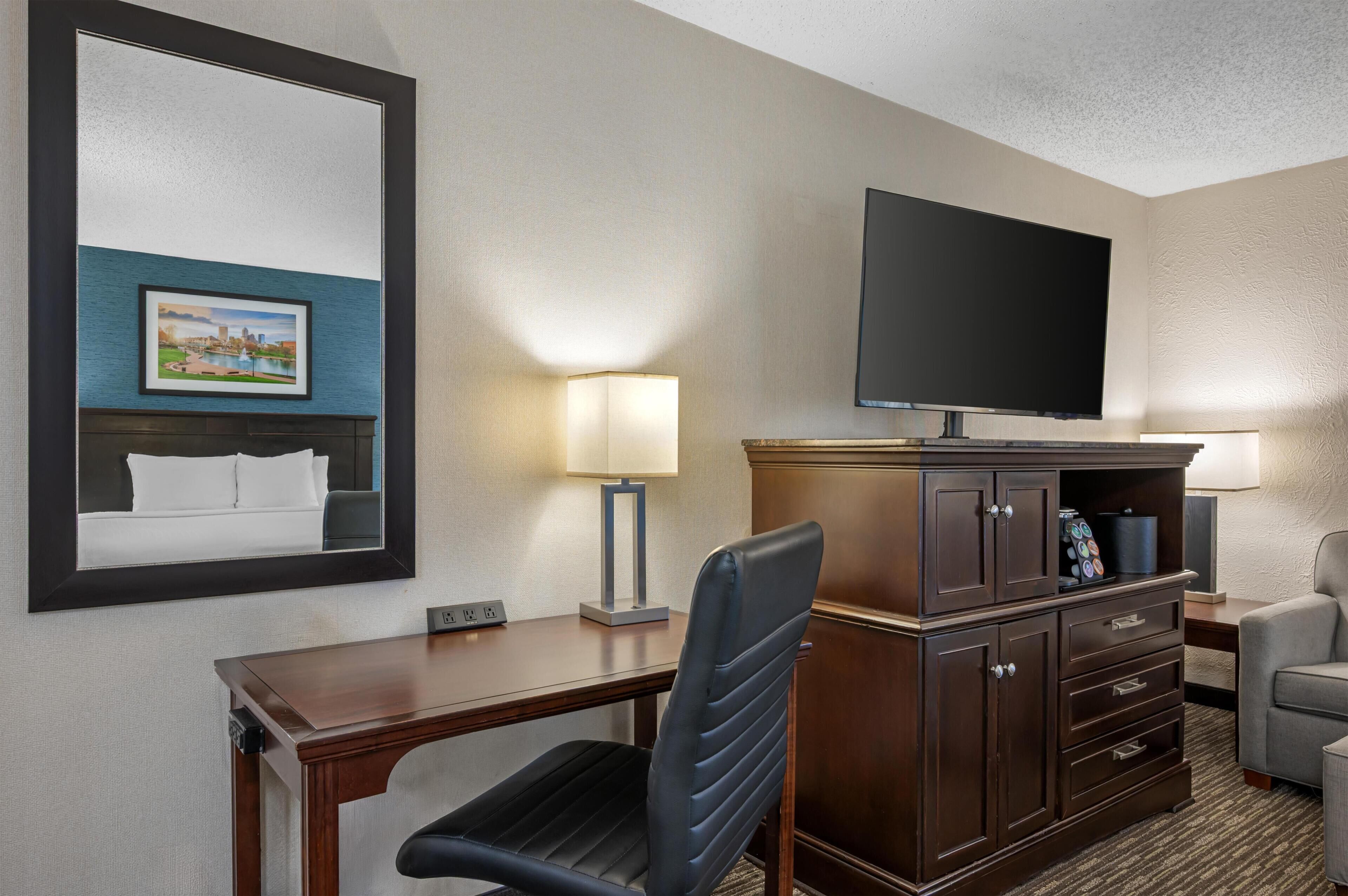 A hotel room with a desk, chair, television, lamp, and a cabinet.
