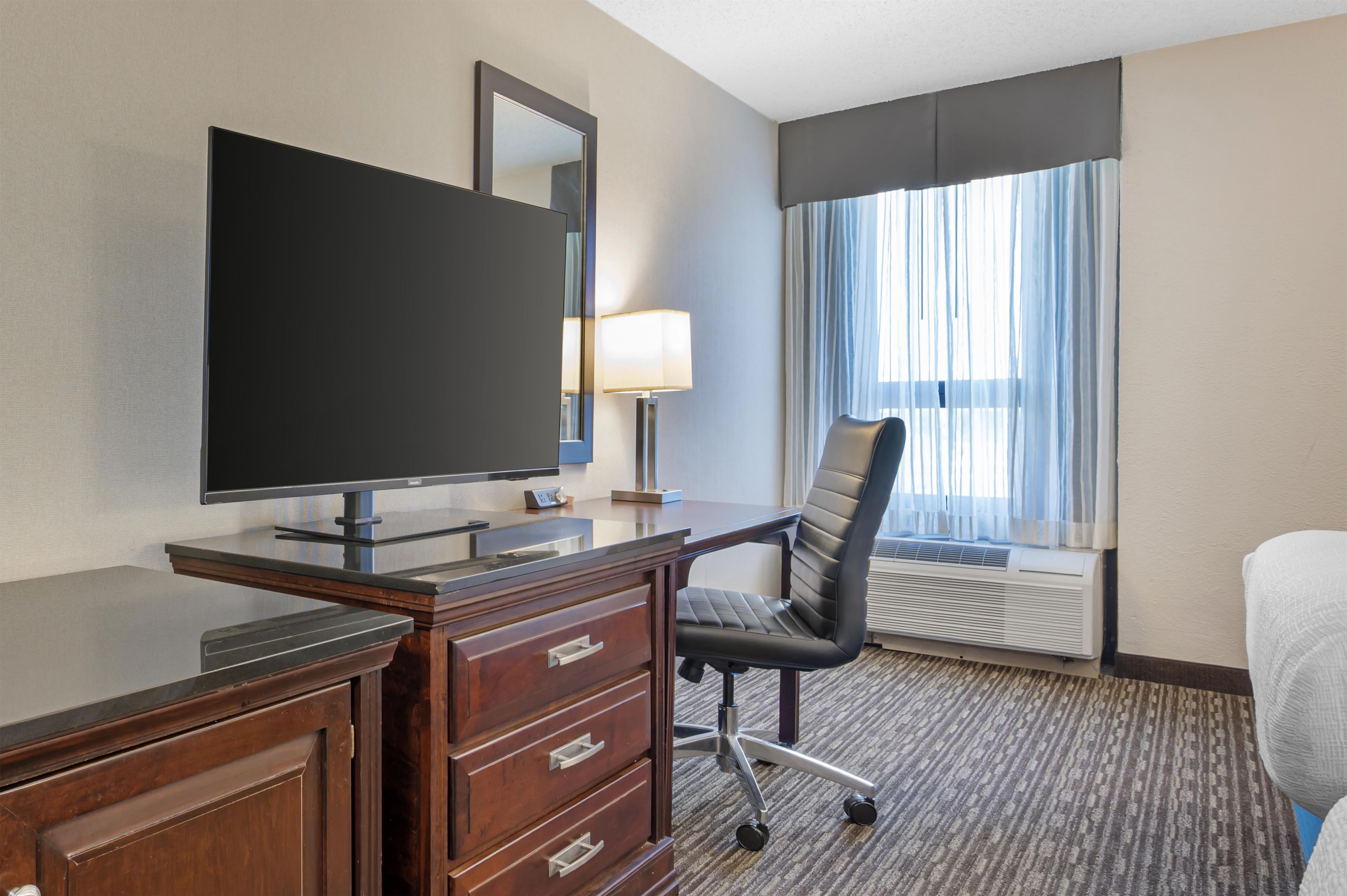 A hotel room with a flat-screen TV, a desk with drawers, a chair, a window with curtains, and an air conditioning unit.