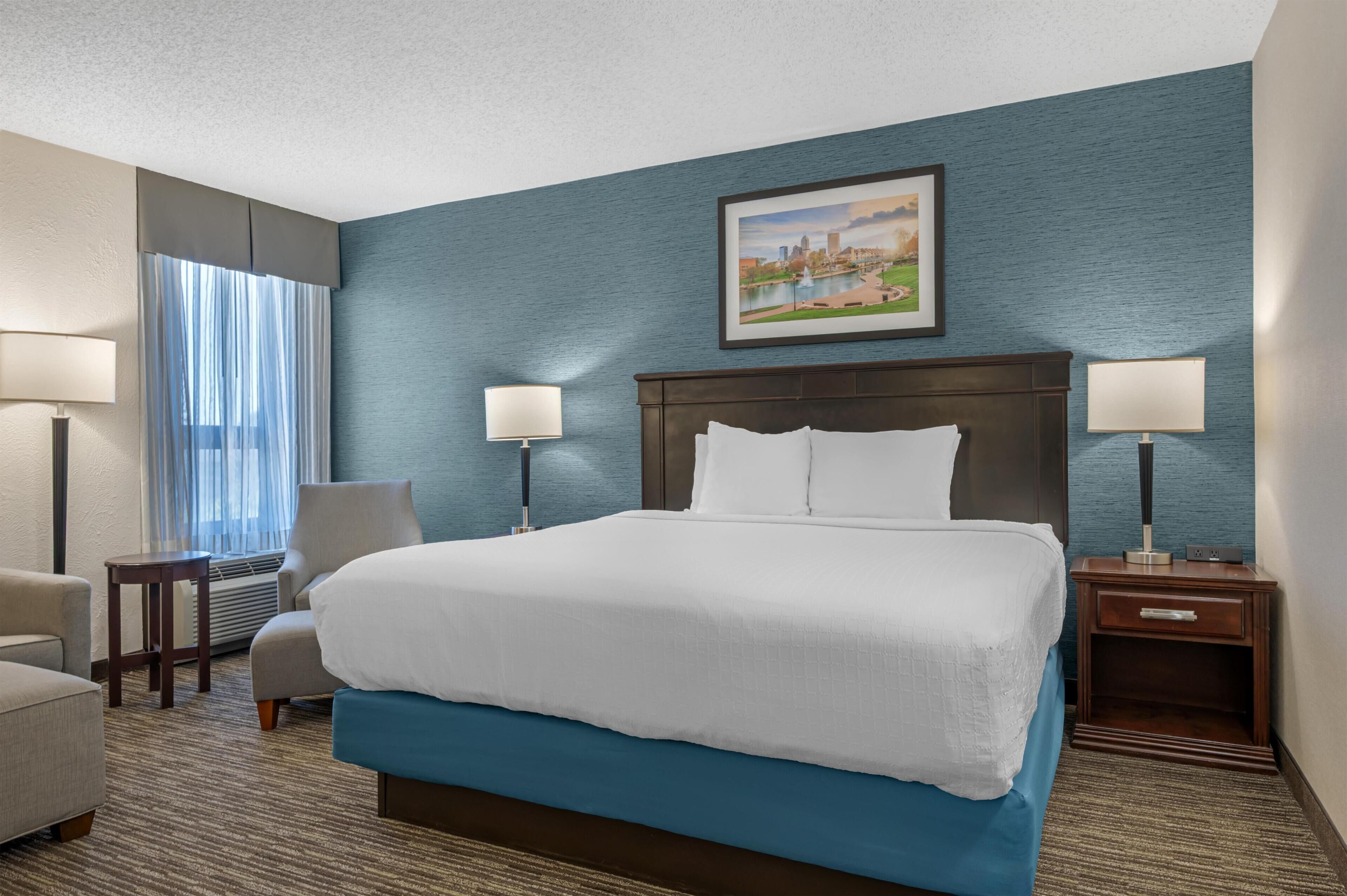 A hotel room with a large bed, two bedside tables with lamps, a chair, and a window with curtains.