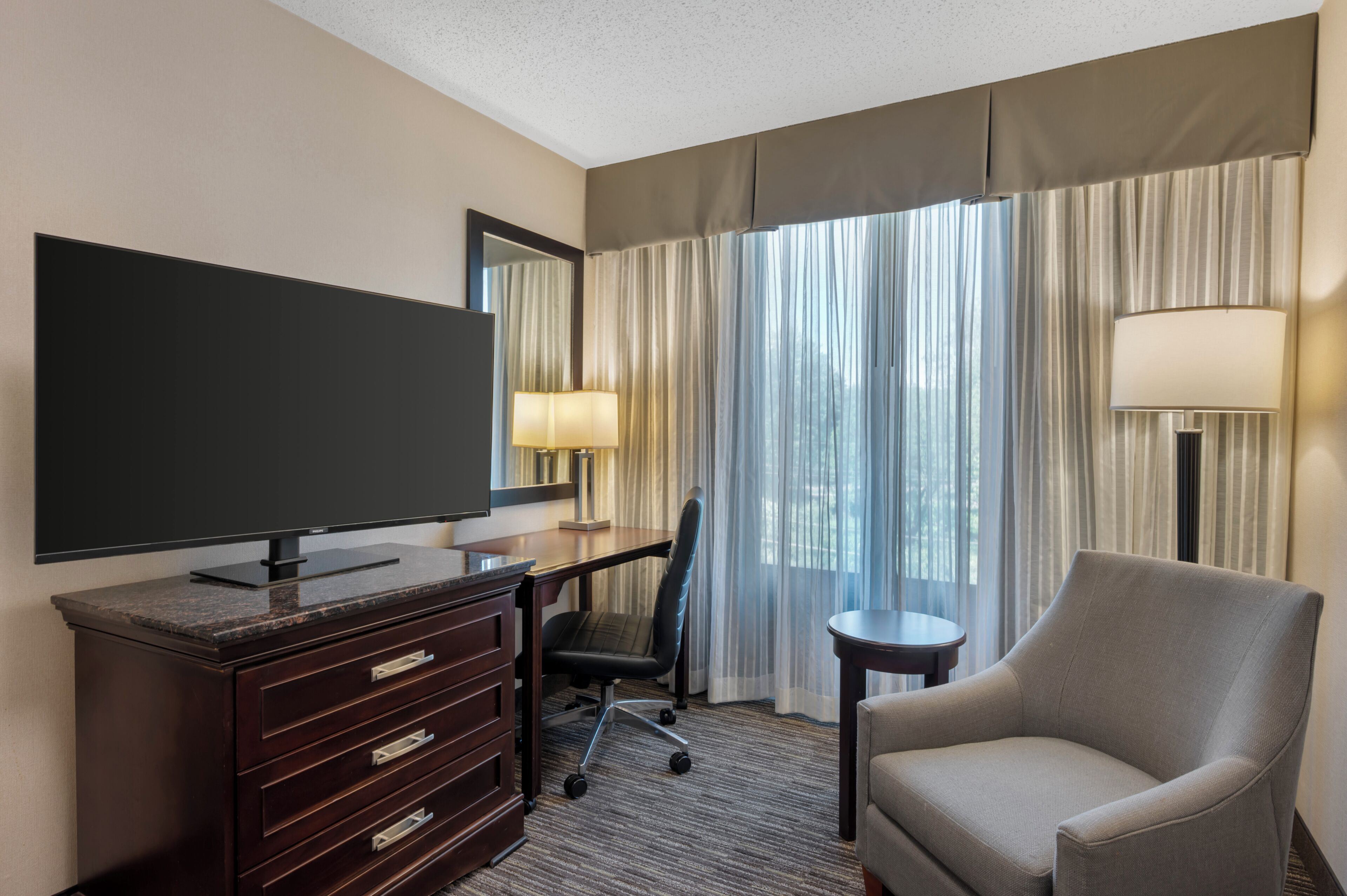 A hotel room with a flat-screen TV, a desk with a chair, a sofa, a lamp, and a large window with curtains.