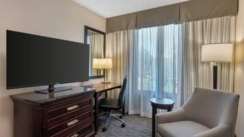 A hotel room with a flat-screen TV, a desk with a chair, a sofa, a lamp, and a large window with curtains.