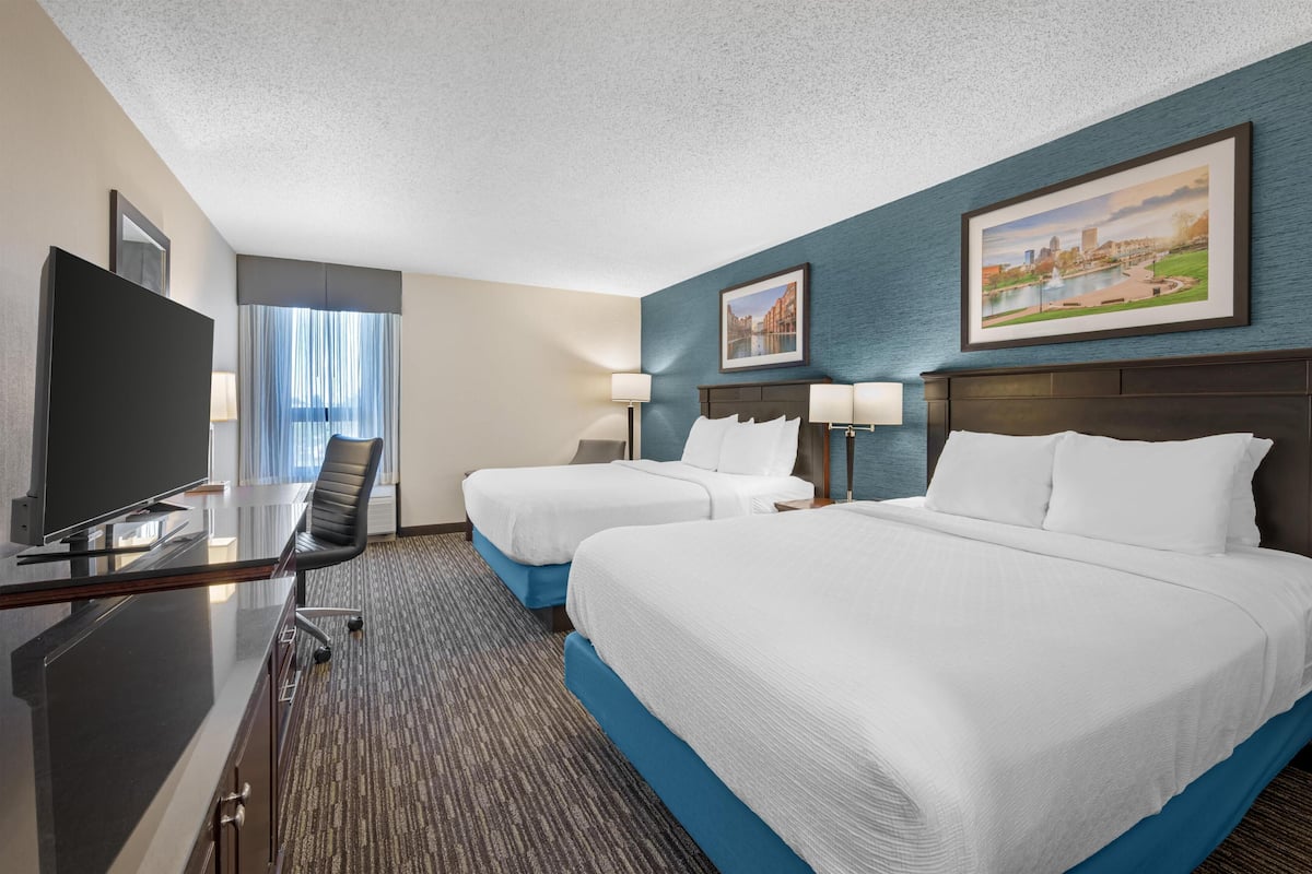 A hotel room with two beds, a desk with a TV, and a view of a cityscape through the window.