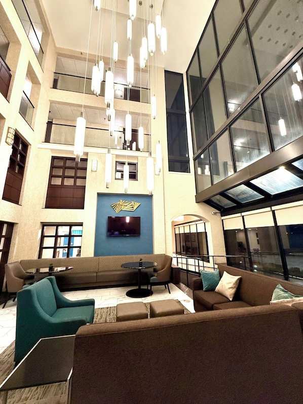 A spacious lobby with a high ceiling, modern pendant lighting, a large flat-screen TV, and a seating area with sofas and a coffee table.