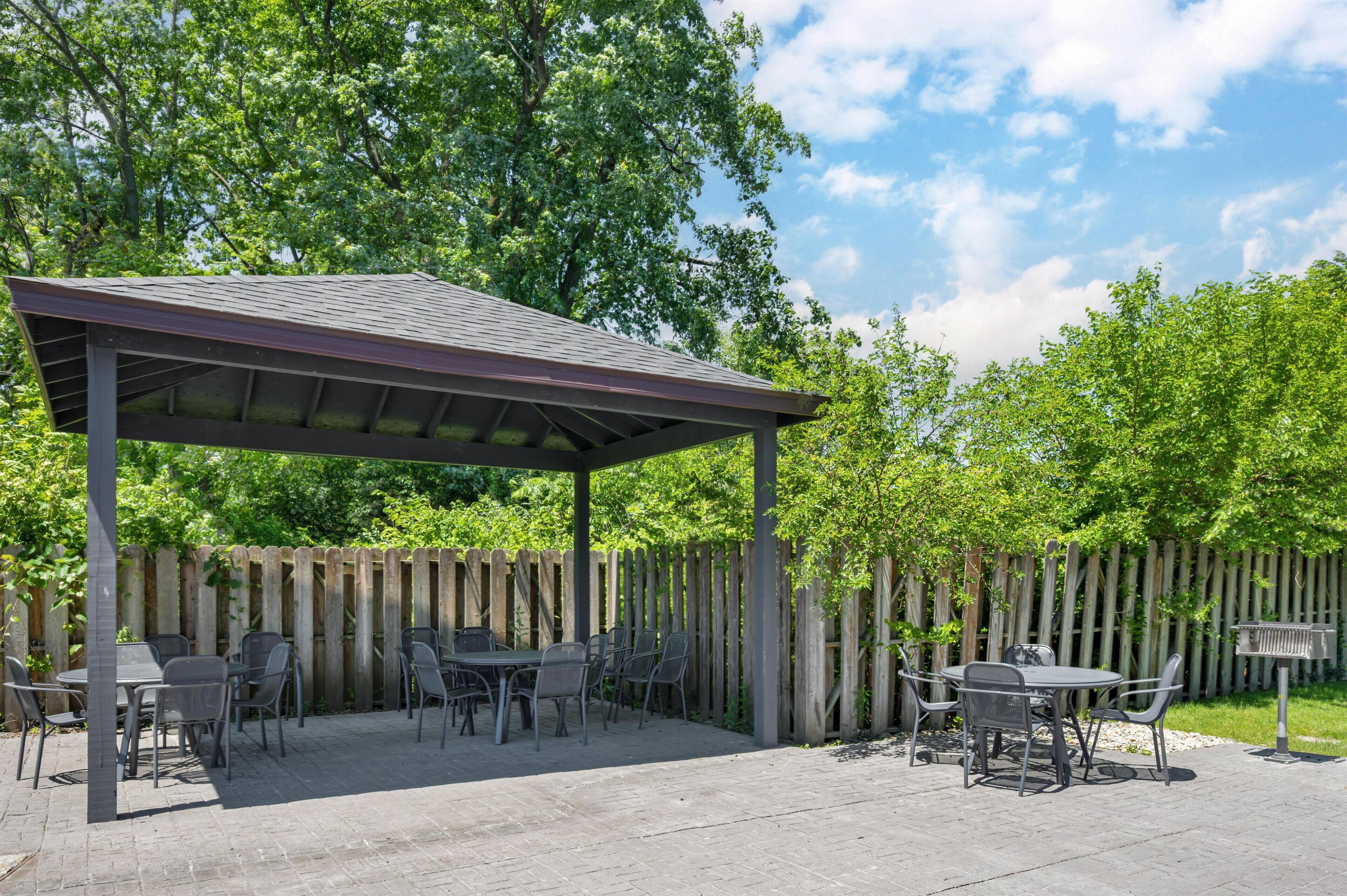Eine überdachte Terrasse mit Tischen und Stühlen, ein Holzzaun und Bäume im Hintergrund.