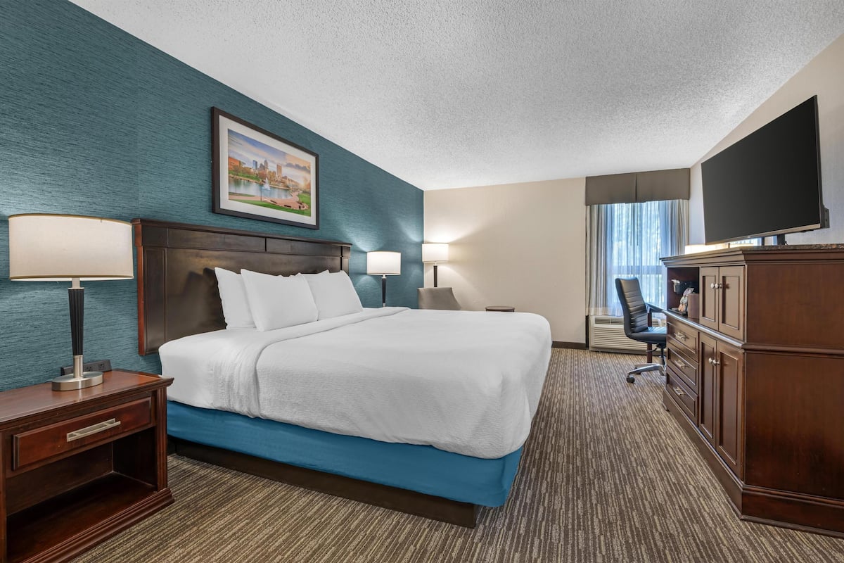 A hotel room with a bed, two bedside tables with lamps, a desk with a chair, a TV, and a large window with curtains.