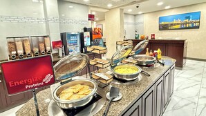En buffetafdeling med skiltede madstationer for proteiner, korn og mejeriprodukter, en vask samt et fjernsyn, der viser et bybillede.