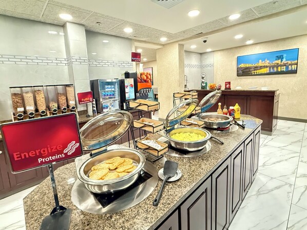 A buffet area with labeled food stations for proteins, grains, and dairy, a sink, and a television displaying a cityscape.