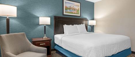A hotel room with a bed, two bedside lamps, a chair, a nightstand, and a framed picture on the wall.