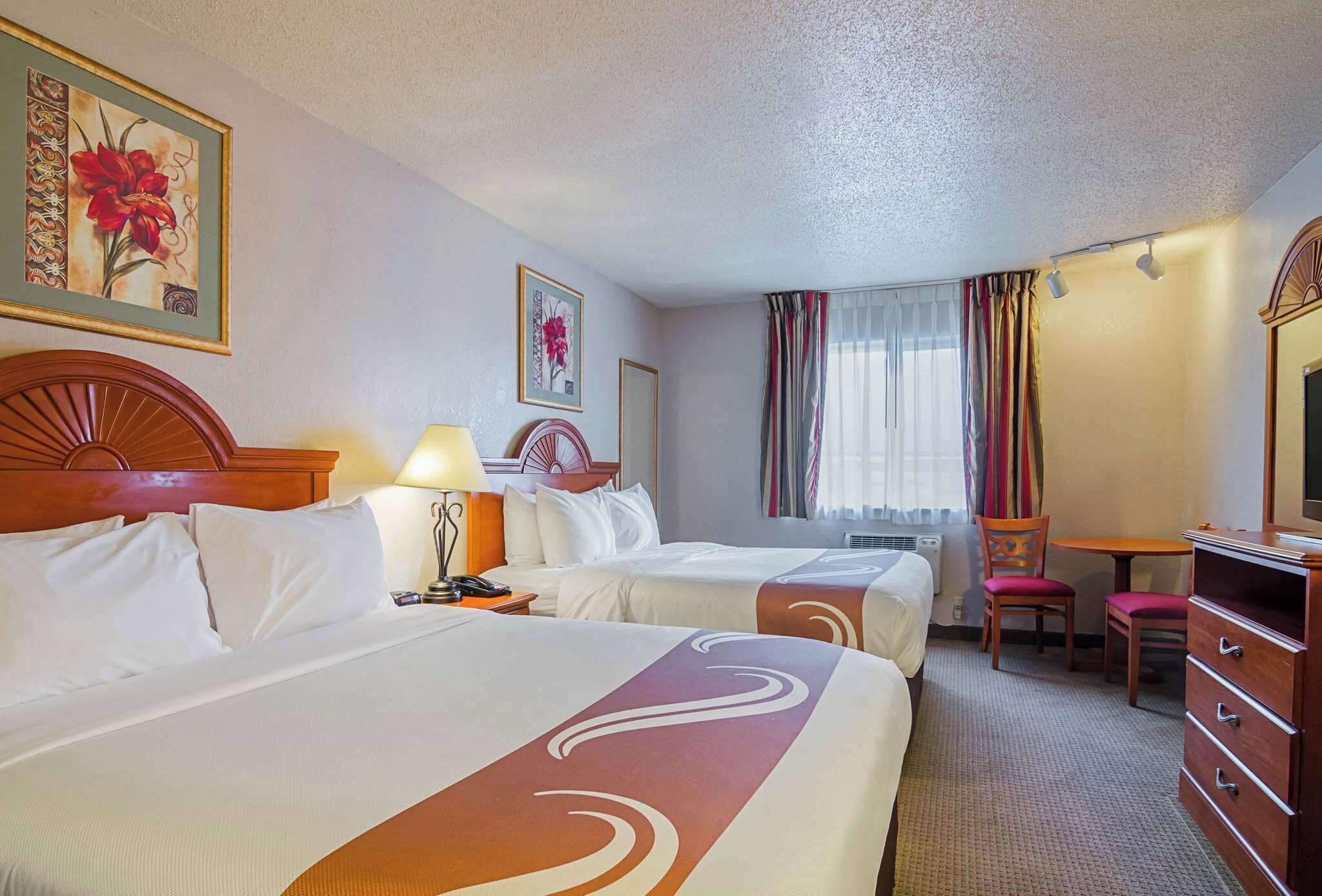 Une chambre d’hôtel avec deux lits, un bureau, une chaise, une commode et une fenêtre avec des rideaux.
