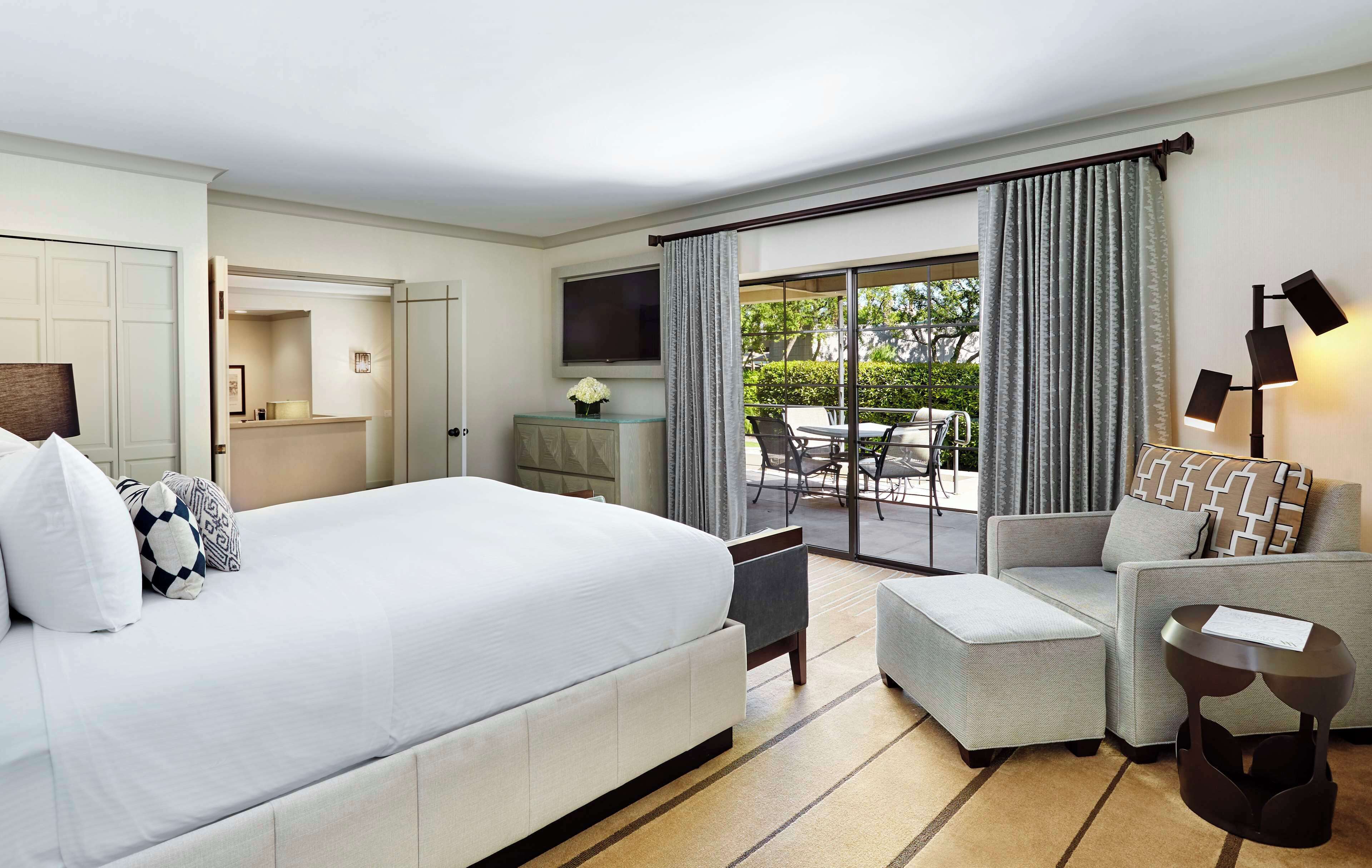A hotel room with a large bed, a sofa, a small table, and a TV. There is a balcony with a table and chairs.