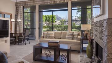 A modern living room with a sofa, coffee table, and fireplace. Large windows offer a view of the outdoors.