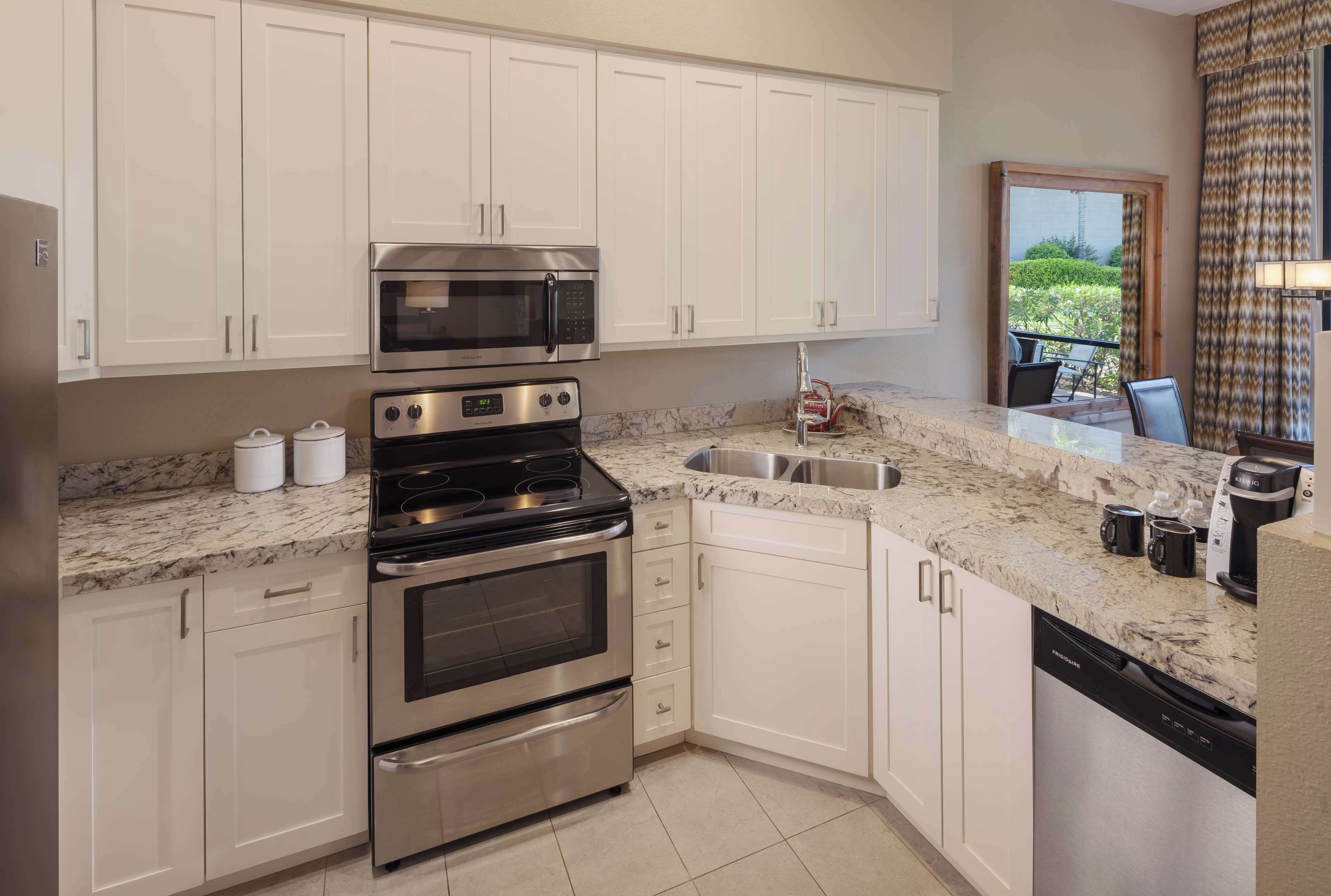 A modern kitchen with white cabinets, stainless steel appliances, a marble countertop, and a view of the outdoors through a sliding door.
