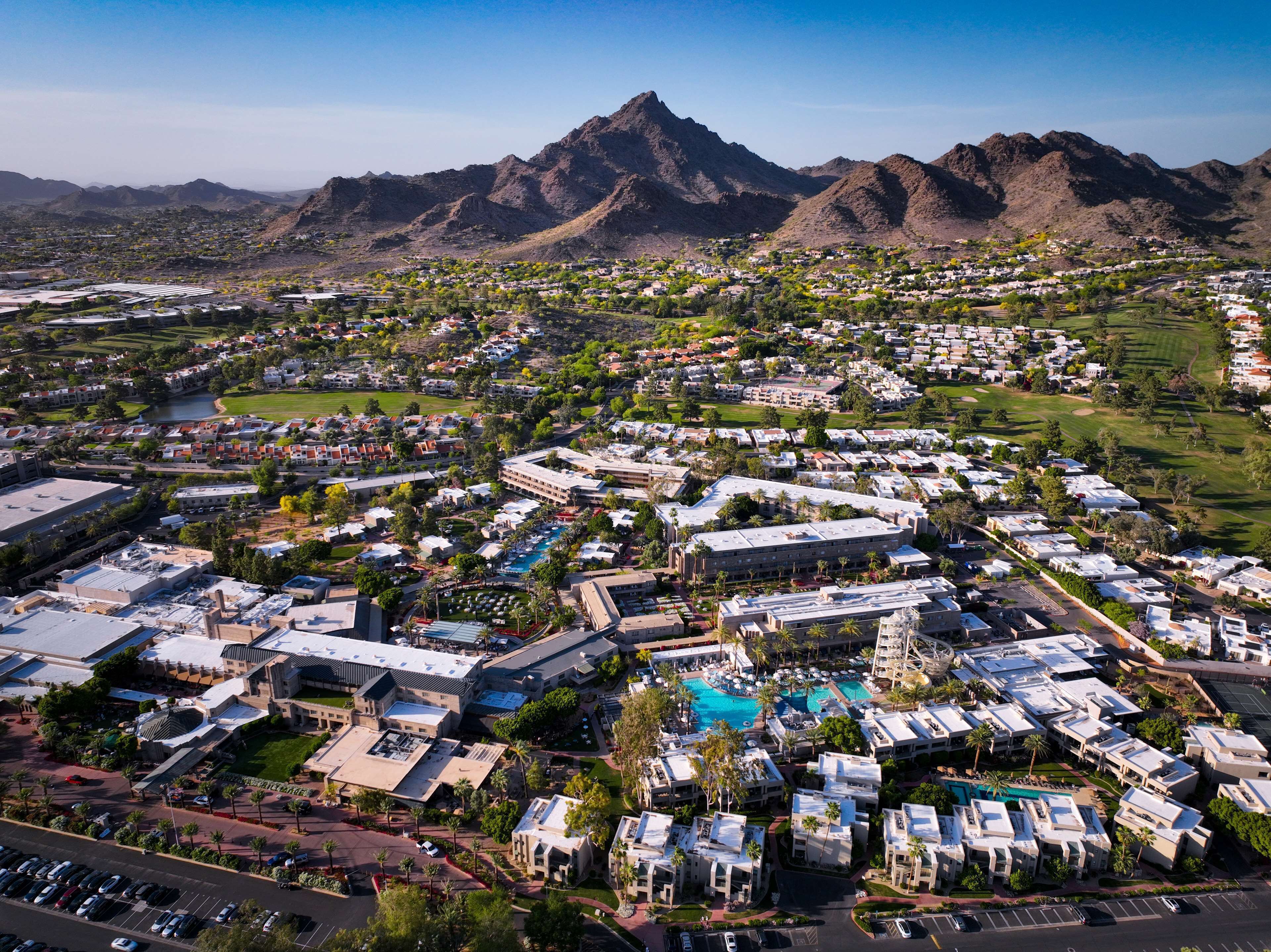An aerial view of a resort area with multiple buildings, a central pool, and a mountainous backdrop.