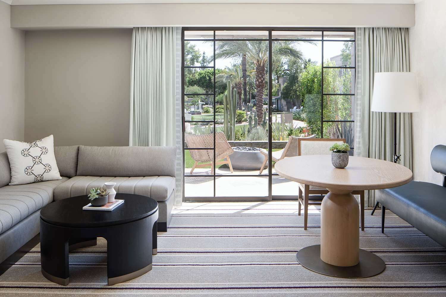 A modern hotel room with a sofa, a round table, and a view of a garden through large windows.