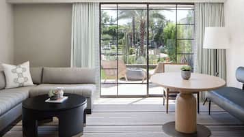 A modern hotel room with a sofa, a round table, and a view of a garden through large windows.