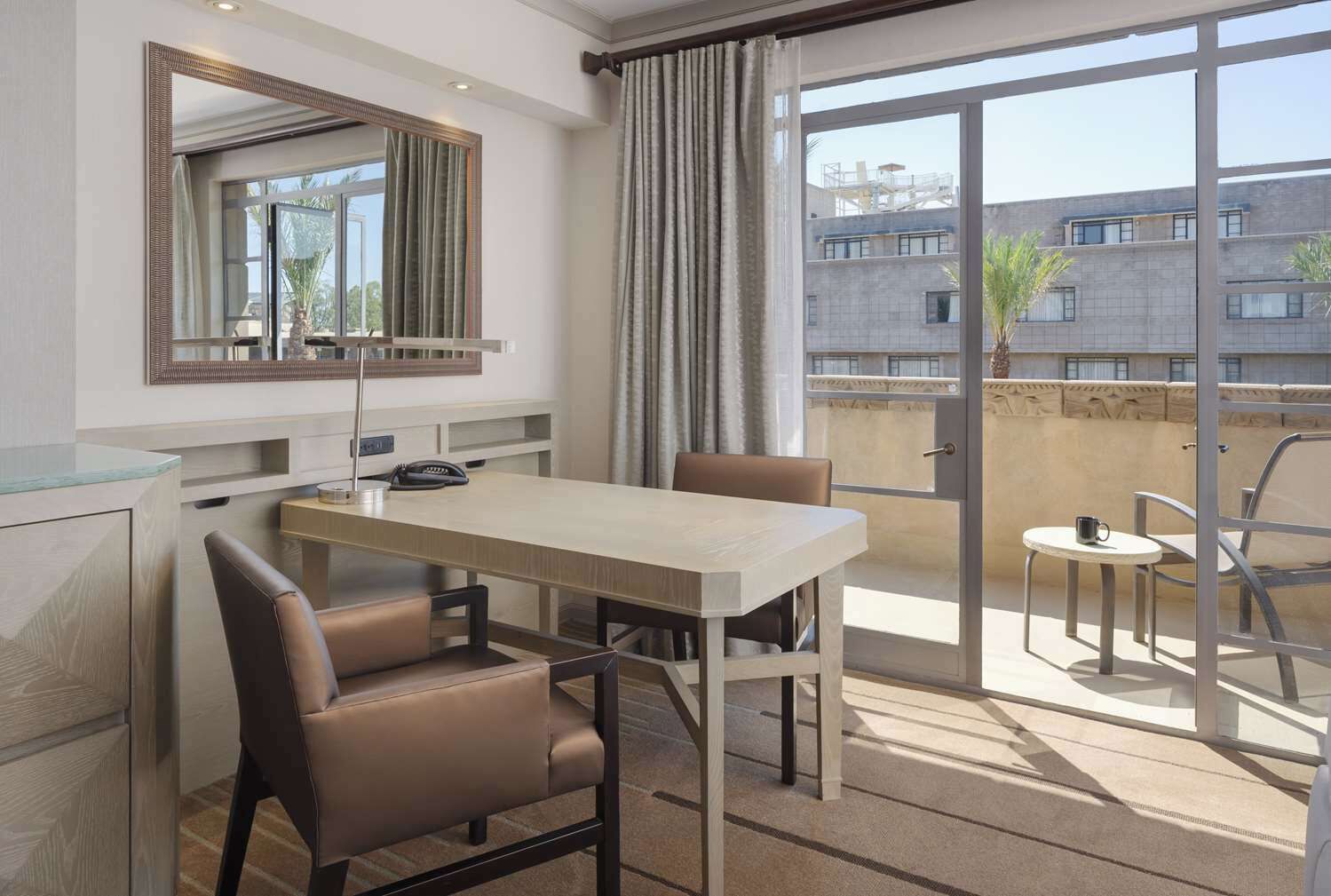 A hotel room with a dining table, two chairs, a large mirror, and a view of the building outside.