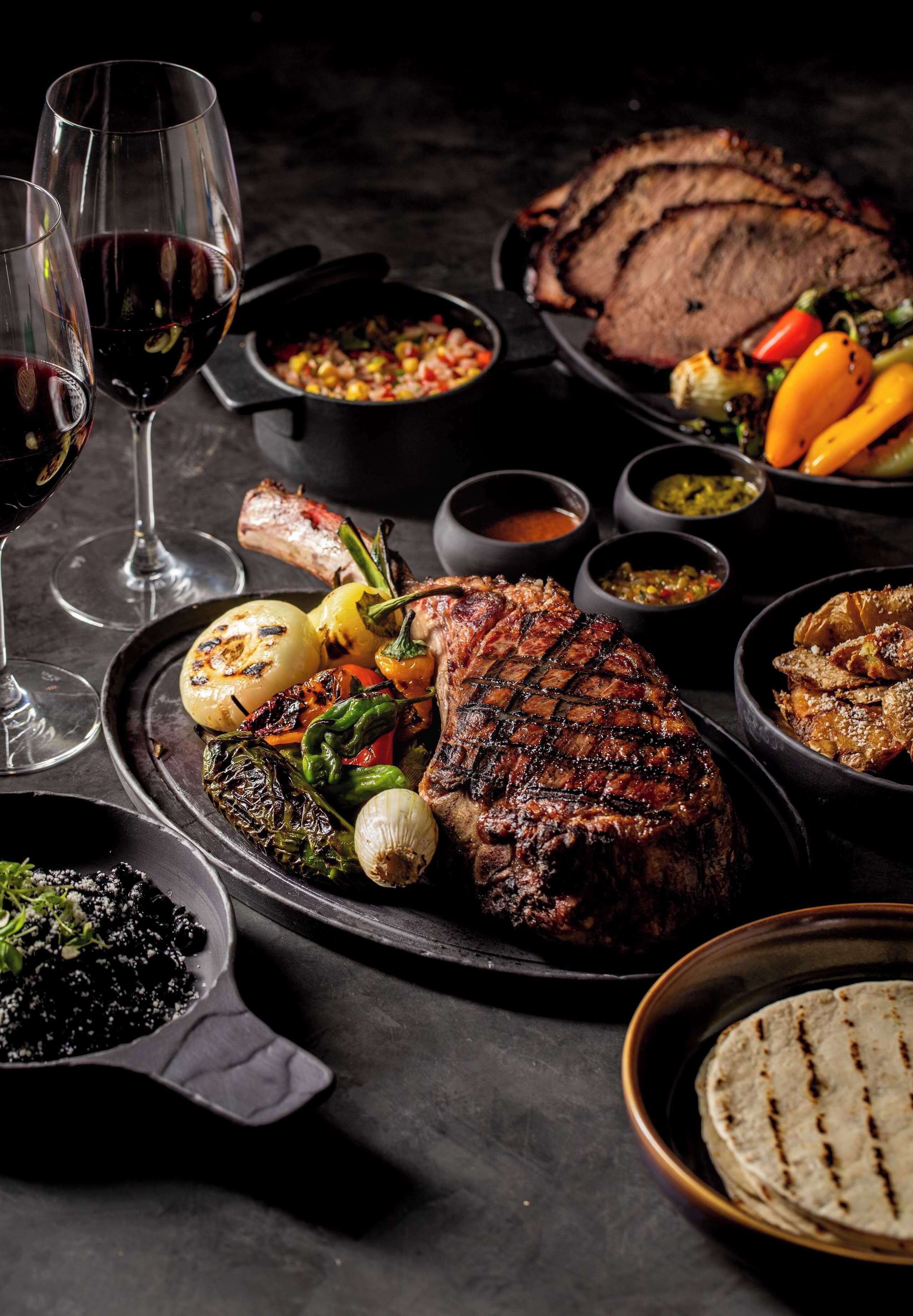 A well-set dining table with a variety of dishes, including grilled meat, vegetables, and sauces, accompanied by wine glasses.