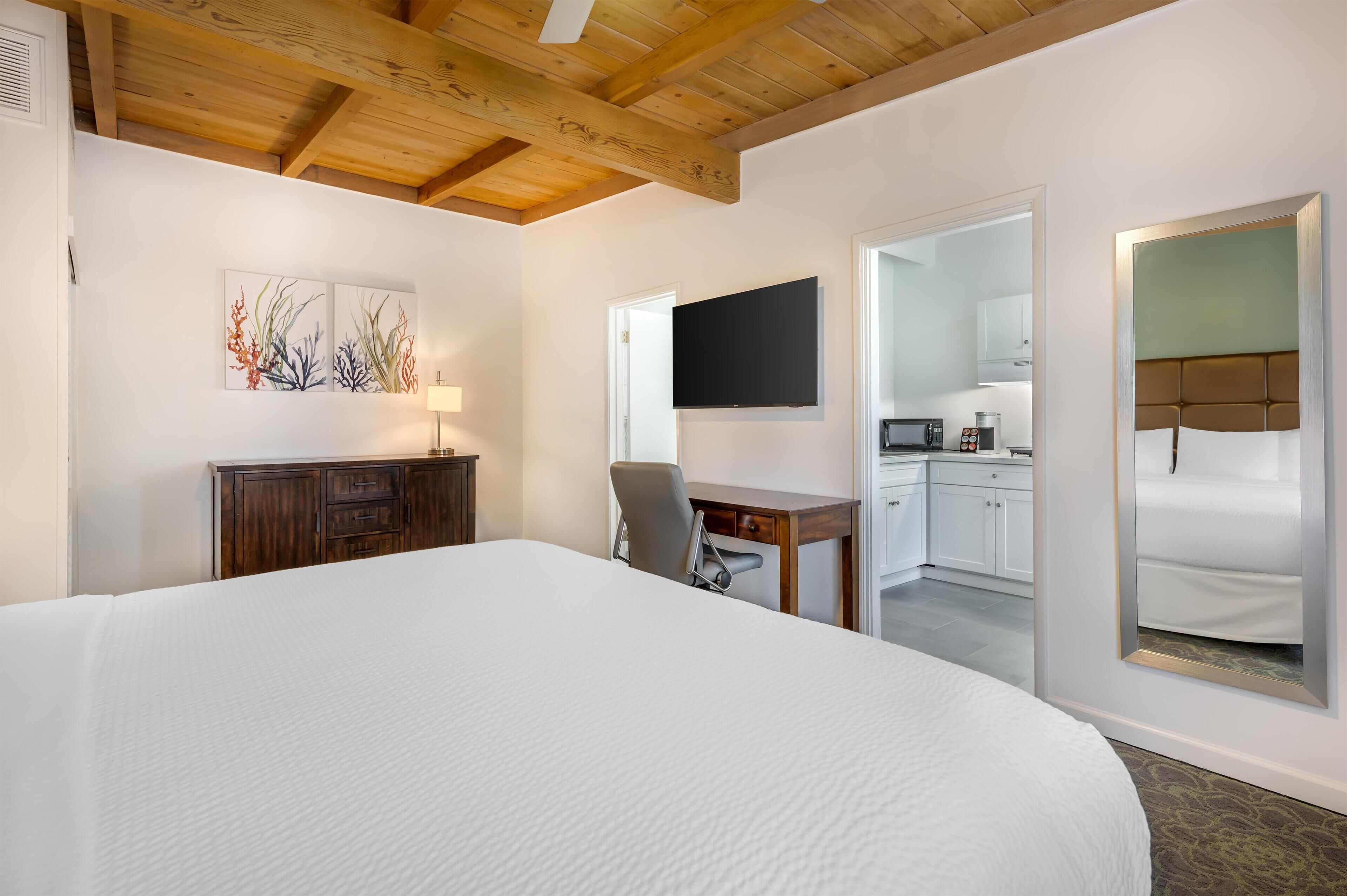 Quarto de hotel com uma cama grande, uma escrivaninha com cadeira, televisão de tela plana e uma área de cozinha visível por uma porta aberta.