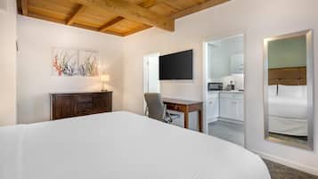 Quarto de hotel com uma cama grande, uma escrivaninha com cadeira, televisão de tela plana e uma área de cozinha visível por uma porta aberta.