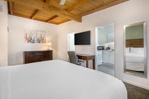 Quarto de hotel com uma cama grande, uma escrivaninha com cadeira, televisão de tela plana e uma área de cozinha visível por uma porta aberta.